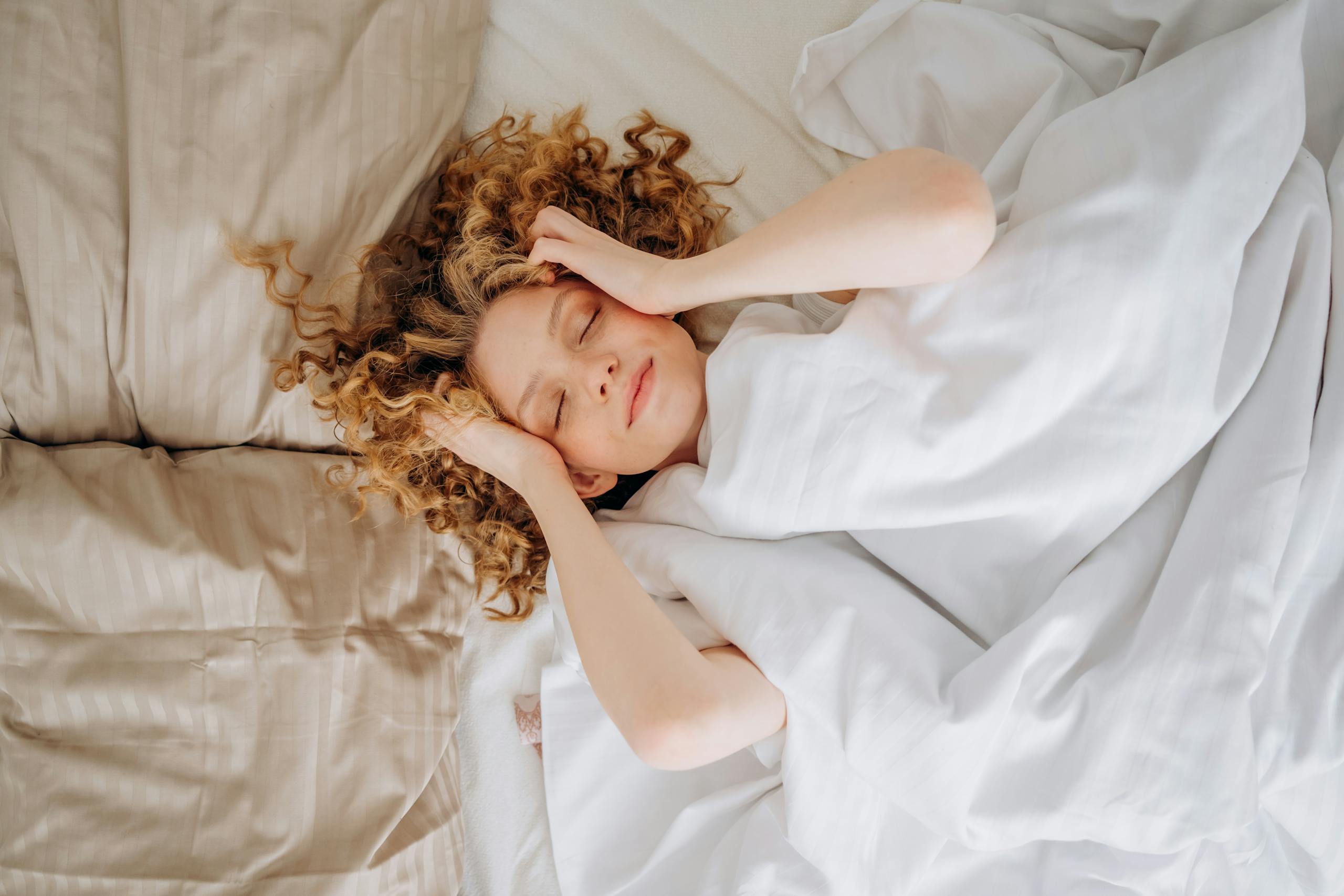 Woman with curly hair sleeping peacefully under white sheets. Indoor comfort.