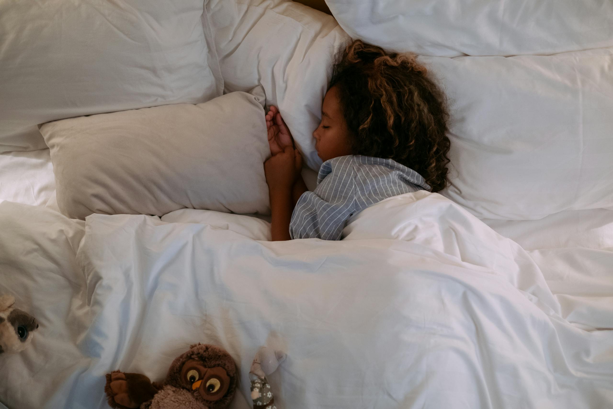 A young girl peacefully sleeping under soft, white bedding with a plush owl nearby.